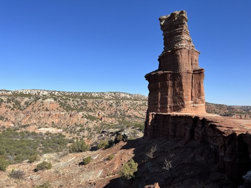 A towering rock formation stands against a clear blue sky, surrounded by rugged terrain and distant hills.