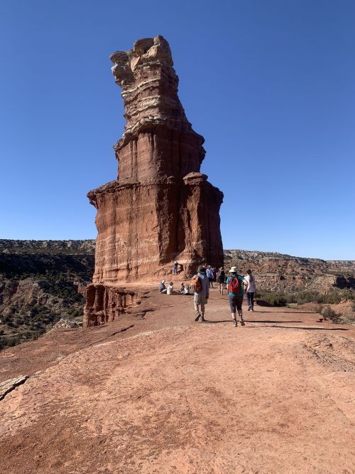 Hikers walking along a rocky path near a tall, unique rock formation under a clear blue sky.