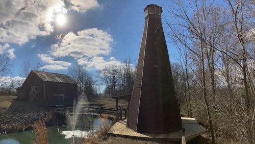 A tall, wooden structure beside a pond, with a barn in the background and a fountain, under a bright, cloudy sky.