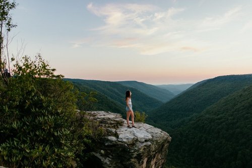 A person stands on a rocky ledge overlooking a lush green valley at sunset, with soft clouds in the sky.