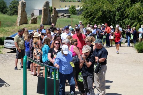 A large crowd of people waiting in line outdoors, with greenery and stone structures in the background.