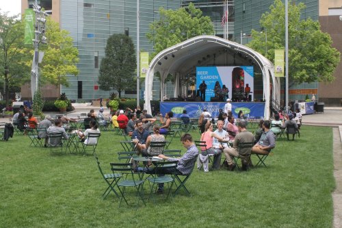 A lively outdoor beer garden with people seated at tables, enjoying drinks and a performance on stage.