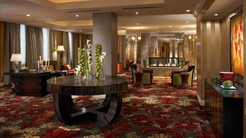 Luxurious hotel lobby with elegant decor, plush seating, and a stylish fireplace, featuring floral carpet and modern lighting.