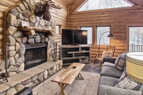 Cozy cabin interior with a stone fireplace, TV, wooden furniture, and large windows overlooking a forest.