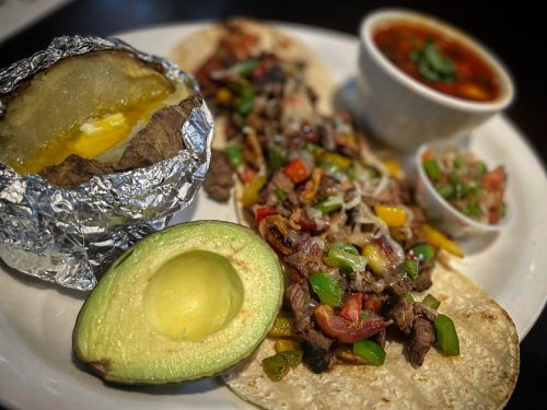A plate featuring tacos with beef, peppers, and toppings, a baked potato, avocado, and a side of salsa.