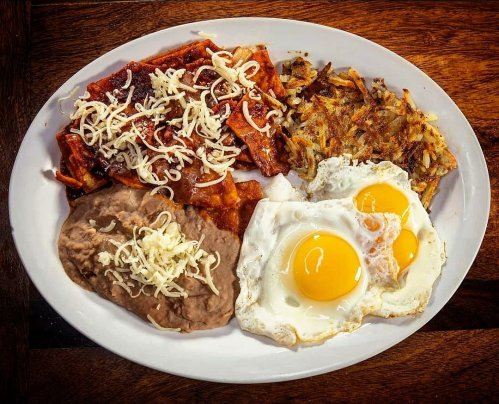 A plate of breakfast featuring enchiladas, refried beans, hash browns, and two sunny-side-up eggs topped with cheese.