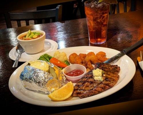 A plate featuring grilled steak with butter, baked potato, fried shrimp, vegetables, and a bowl of soup, alongside a drink.