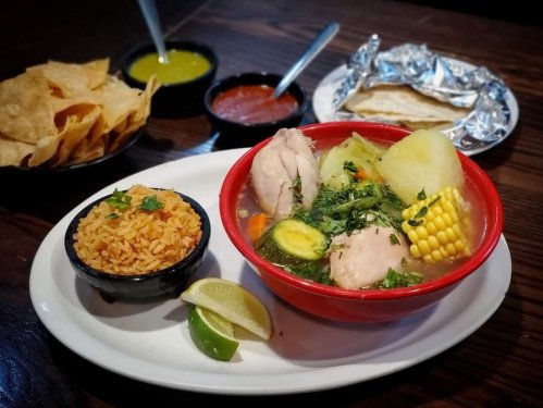 A bowl of chicken soup with vegetables, served with rice, tortillas, and chips on the side.