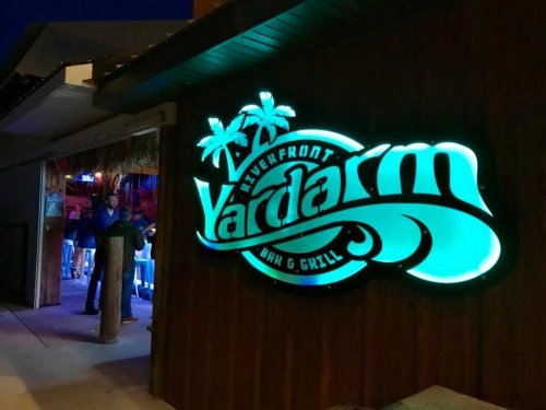 Neon sign for "Vardarm Riverfront Bar & Grill" illuminated at night, with a tropical theme and palm trees.