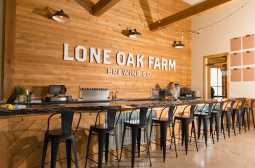 A rustic brewery interior with a wooden bar, stools, and a sign reading "Lone Oak Farm Brewing Co." in a bright space.