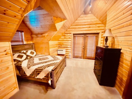 Cozy attic bedroom with wooden walls, a patterned bedspread, and a dresser, featuring sloped ceilings and natural light.