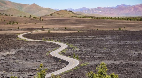 A winding road cuts through a barren landscape of dark lava rock, with mountains in the background under a clear sky.