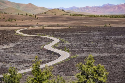 A winding road cuts through a barren landscape of dark lava rock, with mountains in the background and sparse greenery.
