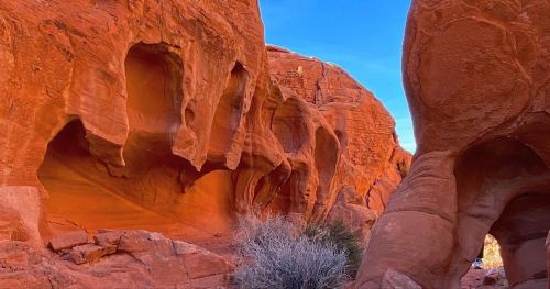 A vibrant red rock formation with unique shapes and textures under a clear blue sky. Sparse vegetation is visible.