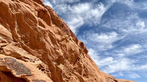 A rugged red rock formation under a blue sky with wispy clouds, showcasing natural textures and patterns.