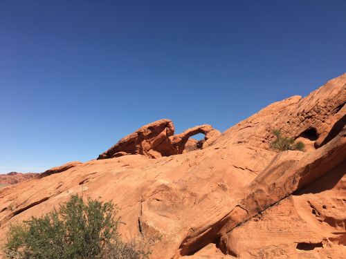 A rocky landscape featuring a natural arch against a clear blue sky. Red sandstone formations are prominent.