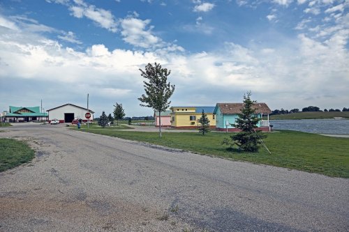 A quiet rural scene featuring colorful buildings, a gravel road, and a pond under a cloudy sky.