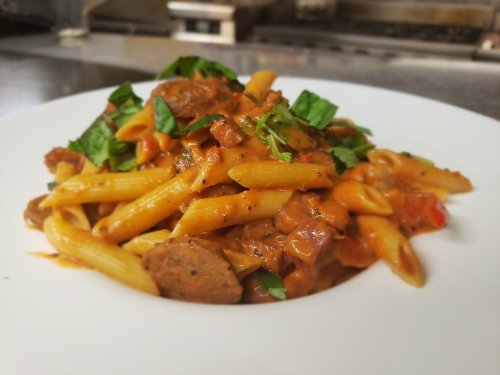 A plate of penne pasta topped with sausage, tomato sauce, and fresh basil, served on a white dish.