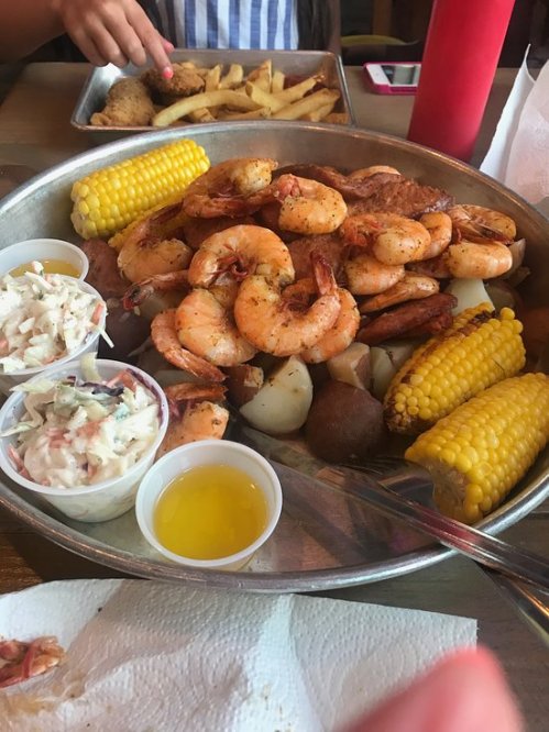 A large metal platter filled with shrimp, corn, potatoes, coleslaw, and dipping sauce, with fries in the background.
