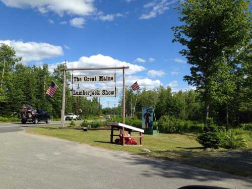 Sign for "The Great Maine Lumberjack Show" with American flags, surrounded by trees and a clear blue sky.