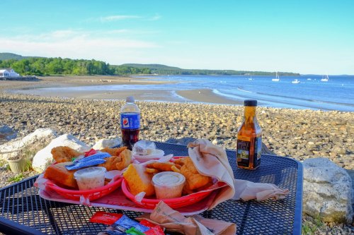 A seaside table with two baskets of fried food, a soda, and a drink, overlooking a calm beach and distant boats.