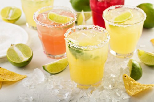 Colorful cocktails with lime slices, garnished with salt, surrounded by lime wedges and tortilla chips on a white surface.