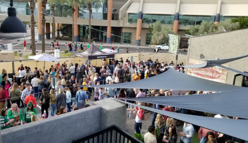 A large crowd gathers at an outdoor event with tents and palm trees, enjoying food and drinks in a lively atmosphere.