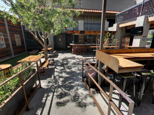 Outdoor seating area of a restaurant with wooden tables, greenery, and a bar in the background.