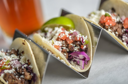 Three tacos filled with meat, diced tomatoes, cheese, and cabbage, served with a lime wedge and a drink in the background.