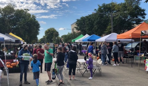 A bustling outdoor market with colorful tents, people shopping, and pumpkins displayed on the ground under a blue sky.