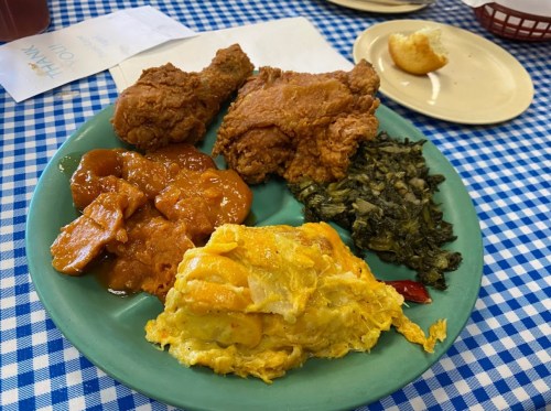 A plate of fried chicken, sweet potatoes, collard greens, and an omelet, served on a blue checkered tablecloth.