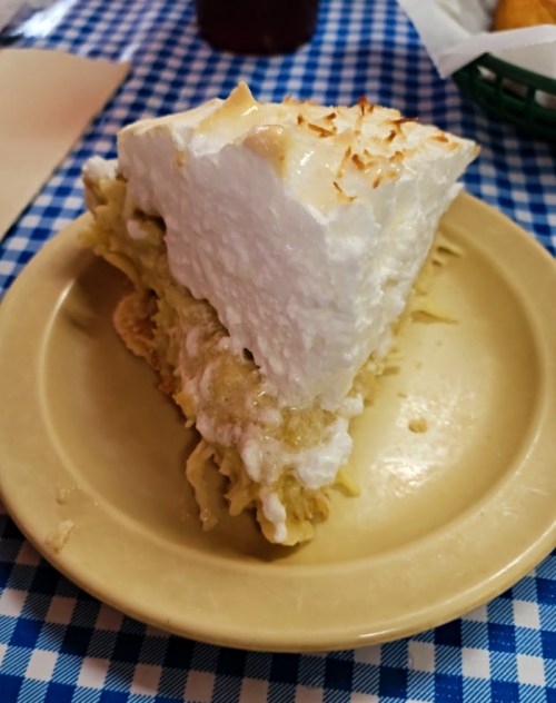 A slice of coconut cream pie on a beige plate, resting on a blue checkered tablecloth.