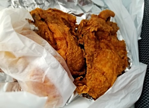 Two pieces of crispy fried chicken in a takeout container, wrapped in white paper and foil.