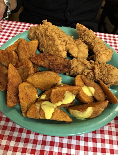 A turquoise plate with crispy fried chicken strips and golden potato wedges drizzled with sauce, on a checkered tablecloth.