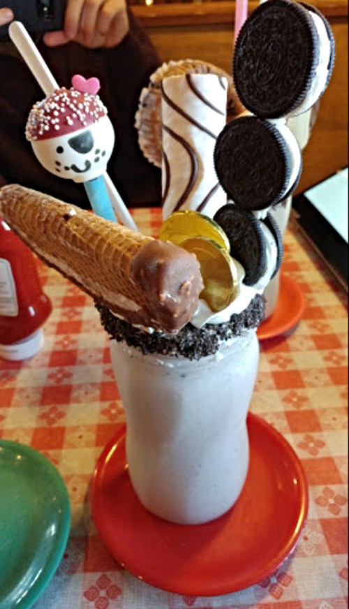 A tall milkshake topped with cookies, a cone, a chocolate-covered treat, and a colorful lollipop on a red plate.