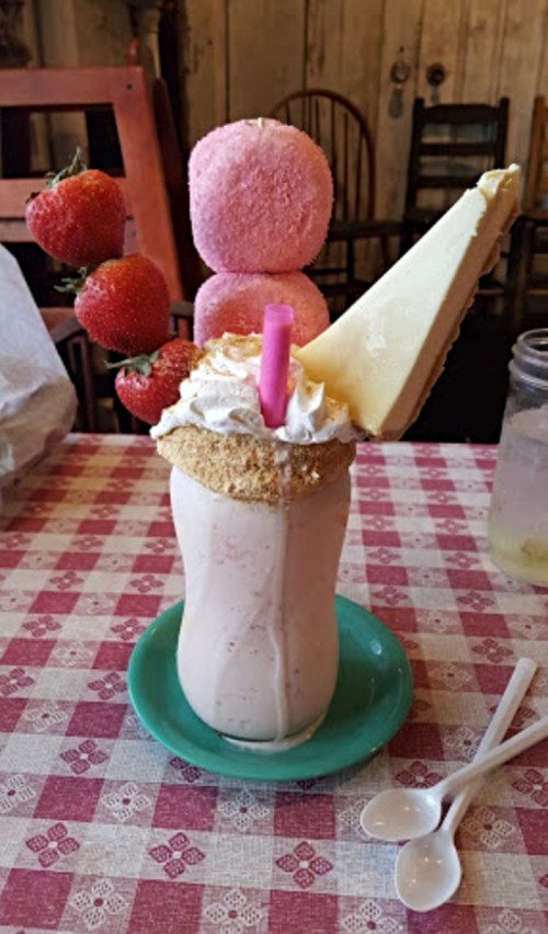 A tall milkshake topped with whipped cream, a slice of cheesecake, strawberries, and pink marshmallows on a checkered tablecloth.