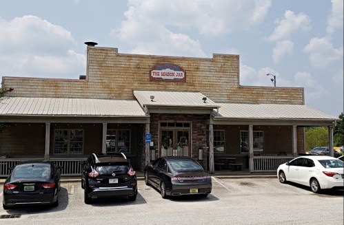 Exterior of a rustic restaurant with a sign reading "The Maison Jar," featuring parked cars in front.