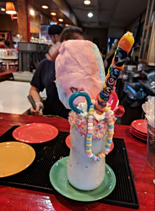 A colorful milkshake topped with cotton candy, candy sticks, and sprinkles, served on a green plate.