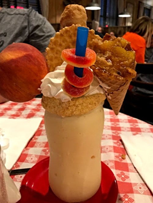 A tall milkshake topped with whipped cream, a cone, gummy rings, and a cookie, served on a red plate.