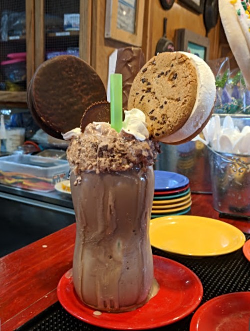 A large chocolate milkshake topped with whipped cream, cookies, and a green straw, served in a colorful diner setting.