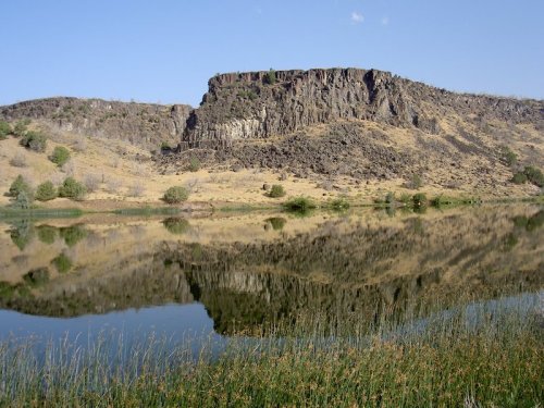 A serene landscape featuring a rocky cliff reflected in a calm lake, surrounded by dry grass and sparse trees.