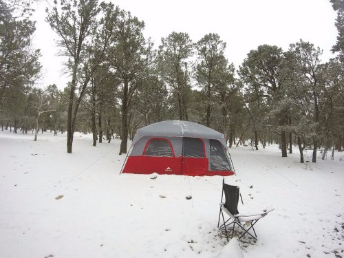 A red and gray tent set up in a snowy forest, with a camping chair nearby among the trees.