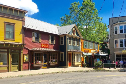 Colorful storefronts line a sunny street, with trees and blue skies in the background. People sit outside at cafes.
