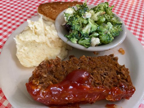 A plate with meatloaf topped with ketchup, mashed potatoes, a side salad, and a slice of toasted bread on a checkered tablecloth.