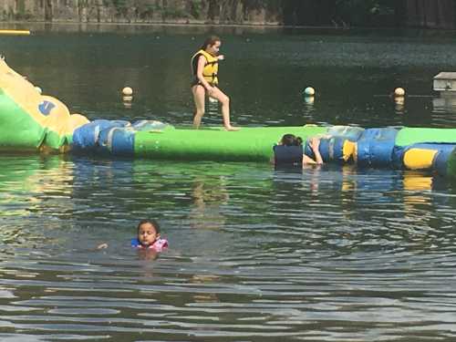 Children playing on a floating obstacle course in a lake, with one girl swimming nearby.