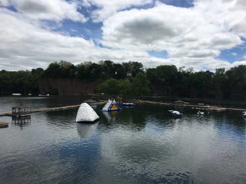 A calm lake with inflatable water structures and a cloudy sky, surrounded by trees and cliffs in the background.