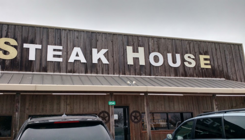 Signage of a rustic steakhouse with large letters on a wooden facade, featuring parked cars in front.