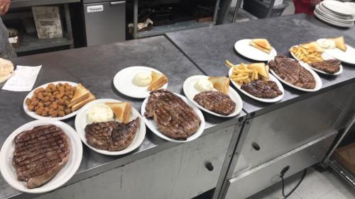 A row of plates with grilled steaks, mashed potatoes, toast, fries, and fried appetizers in a kitchen setting.
