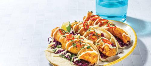 A plate of crispy tacos topped with sliced peppers, cilantro, and drizzled with sauce, served with lime and cabbage.