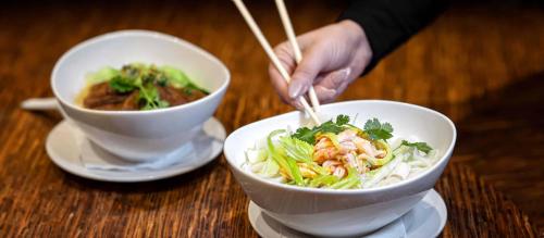 A hand using chopsticks to pick up shrimp and vegetables from a bowl, with another bowl of food in the background.